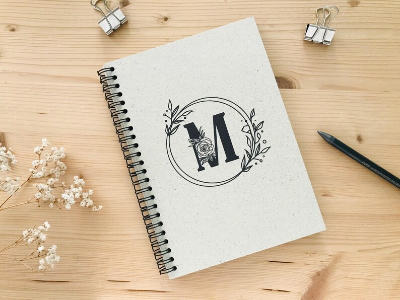 K&ouml;nnte beinhalten: Ein wei&szlig;es Spiralnotizbuch mit einem schwarzen floralen Design und dem Buchstaben "M" in der Mitte. Das Notizbuch hat eine schwarze Spiralbindung.