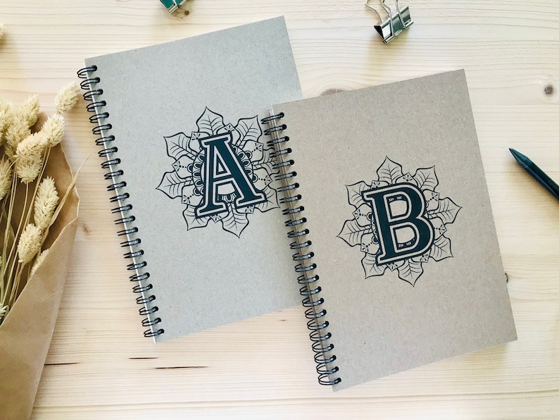 Puede incluir: Dos cuadernos de espiral con cubiertas marrones. Las cubiertas presentan dise&ntilde;os de mandalas en blanco y negro con las letras A y B en el centro.