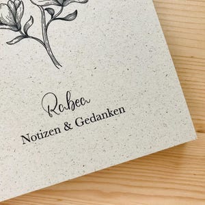 May include: A spiral-bound notebook with a textured, off-white cover. The cover features a delicate floral illustration and the name "Rabea" in cursive, along with the words "Notizen & Gedanken" in a serif font. The notebook rests on a wooden surface.