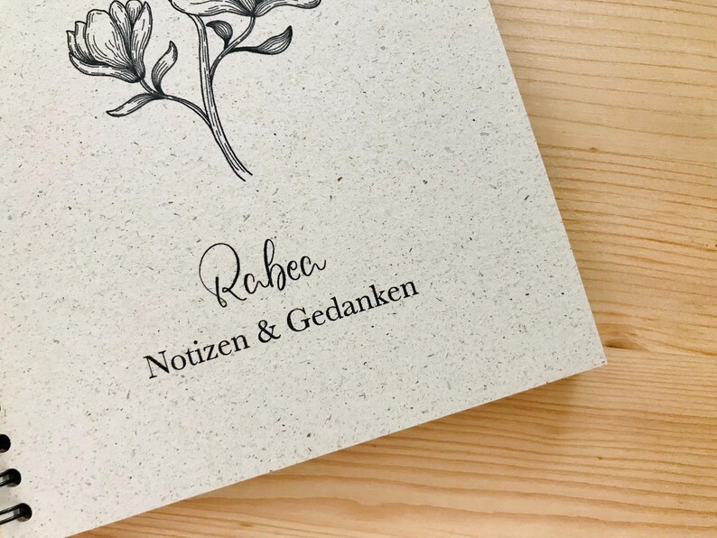 May include: A spiral-bound notebook with a textured, off-white cover. The cover features a delicate floral illustration and the name "Rabea" in cursive, along with the words "Notizen & Gedanken" in a serif font. The notebook rests on a wooden surface.