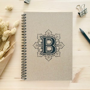 Puede incluir: Un cuaderno marr&oacute;n de espiral con un dise&ntilde;o de mandala negro con la letra "B" en el centro.