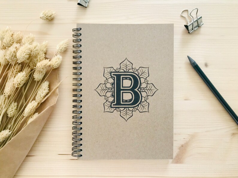 Puede incluir: Un cuaderno marr&oacute;n de espiral con un dise&ntilde;o de mandala negro con la letra "B" en el centro.