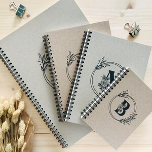 May include: Three brown spiral bound notebooks with black floral designs and the letters A and B on the covers. The notebooks are stacked on top of each other on a wooden surface.