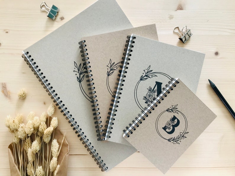 May include: Three brown spiral bound notebooks with black floral designs and the letters A and B on the covers. The notebooks are stacked on top of each other on a wooden surface.