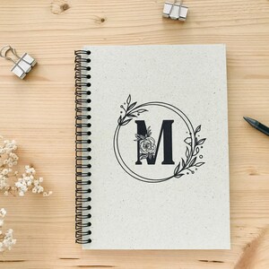 K&ouml;nnte beinhalten: Ein wei&szlig;es Spiralnotizbuch mit einem schwarzen floralen Design und dem Buchstaben "M" in einem Kreis.