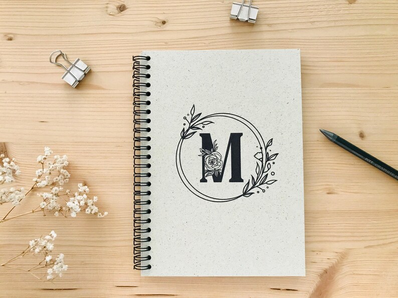 K&ouml;nnte beinhalten: Ein wei&szlig;es Spiralnotizbuch mit einem schwarzen floralen Design und dem Buchstaben "M" in einem Kreis.