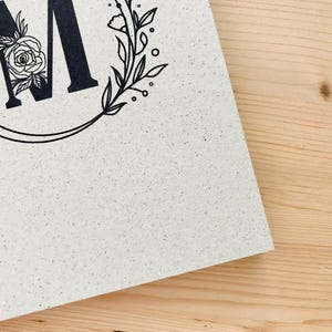 K&ouml;nnte beinhalten: Eine wei&szlig;e Karte mit einem schwarzen floralen Kranz und dem Buchstaben "M" in der Mitte. Der Kranz ist mit kleinen Blumen und Bl&auml;ttern verziert.