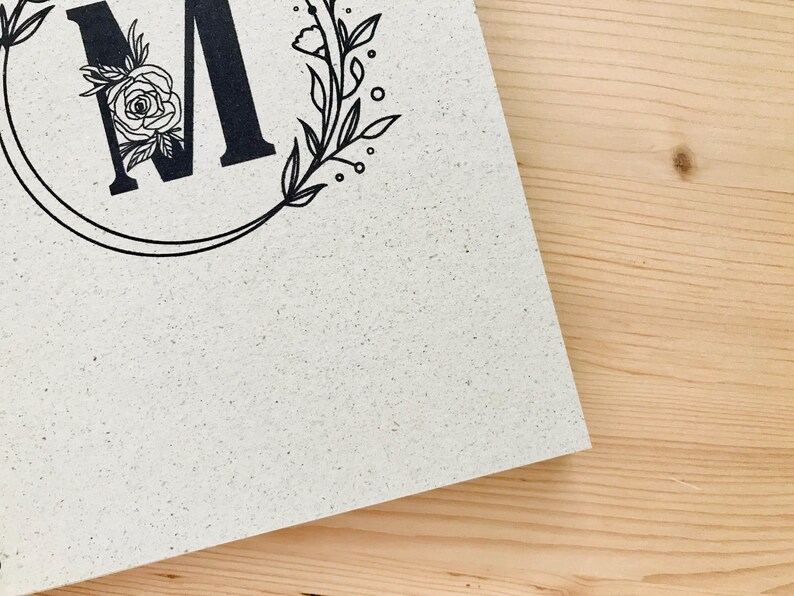 K&ouml;nnte beinhalten: Eine wei&szlig;e Karte mit einem schwarzen floralen Kranz und dem Buchstaben "M" in der Mitte. Der Kranz ist mit kleinen Blumen und Bl&auml;ttern verziert.