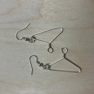May include: A pair of silver-colored wire earrings shaped like miniature clothes hangers. Each earring features a hook and a small, round bead. The earrings are displayed on a light-colored wooden surface.