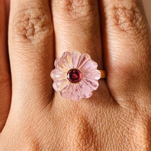 Può includere: Un anello a forma di fiore rosa con un design intagliato e una piccola pietra preziosa rosso scuro al centro.
