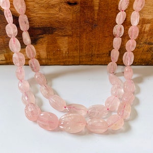 Puede incluir: Un hilo de cuentas de piedras preciosas ovaladas de color rosa. Las cuentas son translúcidas y tienen un aspecto ligeramente nublado. Las cuentas están enhebradas en un hilo transparente.