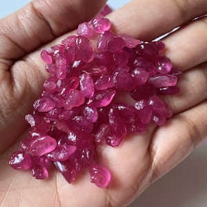 Puede incluir: Un puñado de piedras preciosas de rubí en bruto. Las pequeñas piedras irregulares son de un color rojo rosado intenso, con una calidad translúcida. Las piedras preciosas se muestran en una mano abierta, mostrando su apariencia natural y en bruto.