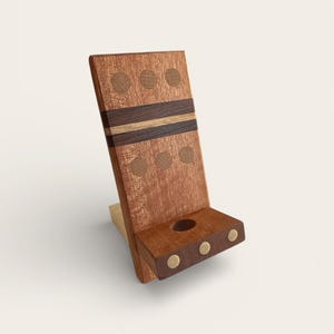 May include: A wooden phone stand with a reddish-brown finish. The stand features decorative inlaid circles and stripes in varying shades of brown. The base has a small shelf with a hole and brass accents.