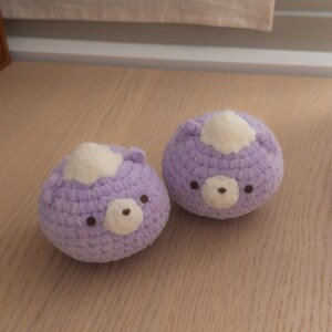 Peut inclure: Deux jouets faits &agrave; la main en forme d'ours au crochet. Chaque ours est de couleur lavande douce avec une tache blanche sur la t&ecirc;te et un petit nez et des yeux bruns. Les jouets sont sur une surface en bois clair.