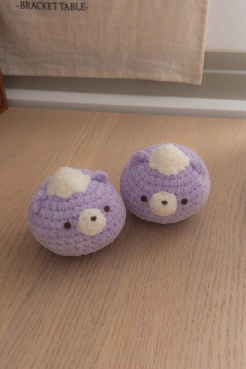 Peut inclure: Deux jouets faits &agrave; la main en forme d'ours au crochet. Chaque ours est de couleur lavande douce avec une tache blanche sur la t&ecirc;te et un petit nez et des yeux bruns. Les jouets sont sur une surface en bois clair.