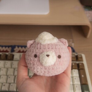 Peut inclure: Un ours rose crochet&eacute; &agrave; la main avec un chapeau blanc et duveteux. L'ours a de petits yeux bruns et un museau blanc. Le jouet en peluche est tenu dans une main, avec un clavier et un bureau en arri&egrave;re-plan.