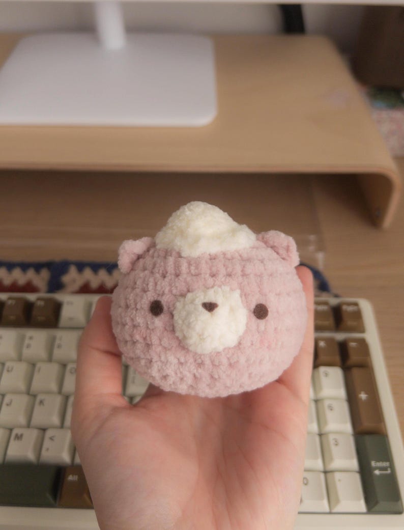 Peut inclure: Un ours rose crochet&eacute; &agrave; la main avec un chapeau blanc et duveteux. L'ours a de petits yeux bruns et un museau blanc. Le jouet en peluche est tenu dans une main, avec un clavier et un bureau en arri&egrave;re-plan.