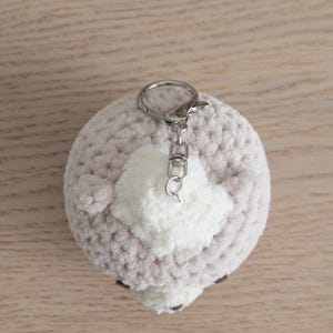 Peut inclure: Un porte-cl&eacute;s rond fait &agrave; la main, avec un corps crochet&eacute; gris clair et un accent en pompon blanc. Le porte-cl&eacute;s a un fermoir argent&eacute; et une petite boucle pour attacher les cl&eacute;s. L'article est pos&eacute; sur une surface en bois brun clair.