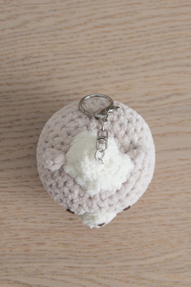 Peut inclure: Un porte-cl&eacute;s rond fait &agrave; la main, avec un corps crochet&eacute; gris clair et un accent en pompon blanc. Le porte-cl&eacute;s a un fermoir argent&eacute; et une petite boucle pour attacher les cl&eacute;s. L'article est pos&eacute; sur une surface en bois brun clair.