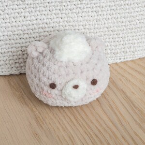 Peut inclure: Un ours en crochet rose clair avec une touffe blanche sur la t&ecirc;te, des yeux bruns et des joues roses. L'ours est pos&eacute; sur une surface en bois, avec un fond en crochet blanc. Ce jouet doux est parfait pour un b&eacute;b&eacute;.