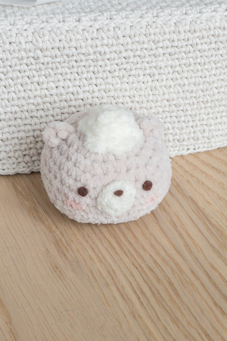 Peut inclure: Un ours en crochet rose clair avec une touffe blanche sur la t&ecirc;te, des yeux bruns et des joues roses. L'ours est pos&eacute; sur une surface en bois, avec un fond en crochet blanc. Ce jouet doux est parfait pour un b&eacute;b&eacute;.