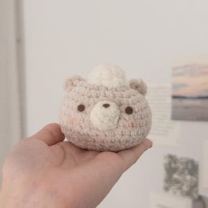 Peut inclure: Un ours en crochet fait &agrave; la main, de couleur beige clair, avec de petits yeux bruns et un dessus blanc et duveteux. L'ours rond est doux, id&eacute;al comme jouet pour enfant ou d&eacute;coration de chambre d'enfant. L'ours est tenu dans une main.