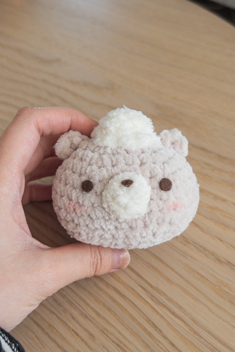 Peut inclure: Un jouet en forme d'ours rond et moelleux, de couleur beige clair, avec un chapeau pompon blanc. L'ours a des yeux bruns et un museau blanc. Des joues roses accentuent le tout, cr&eacute;ant une esth&eacute;tique mignonne et faite &agrave; la main.
