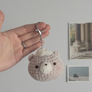 Peut inclure: Un porte-cl&eacute;s ours crochet&eacute; en rose clair et blanc. L'ours a de petits yeux fonc&eacute;s, un museau blanc et des joues roses. Il est attach&eacute; &agrave; un porte-cl&eacute;s de couleur argent.