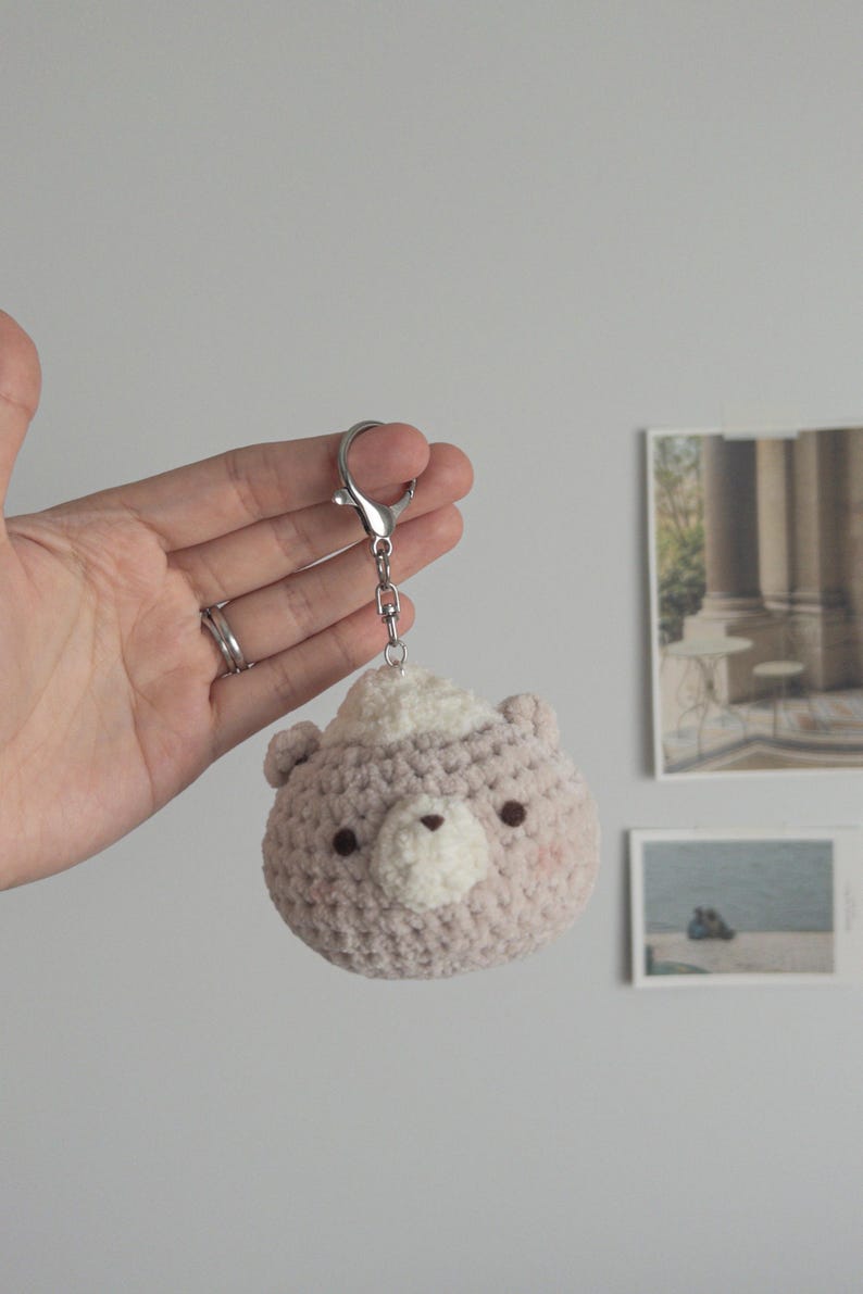 Peut inclure: Un porte-cl&eacute;s ours crochet&eacute; en rose clair et blanc. L'ours a de petits yeux fonc&eacute;s, un museau blanc et des joues roses. Il est attach&eacute; &agrave; un porte-cl&eacute;s de couleur argent.
