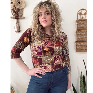 May include: A long-sleeve top featuring a patchwork design with various floral and ornate patterns in shades of burgundy, gold, and brown. The garment has a round neckline and three-quarter length sleeves with a subtle lettuce-edge hem finish.