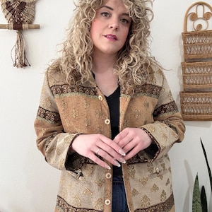 Puede incluir: Chaqueta estampada marrón y beige con botones. La chaqueta presenta un diseño floral y se lleva sobre una camiseta negra y vaqueros azules. La chaqueta tiene una estética vintage.