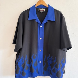 May include: Black button-up shirt with a blue collar and flame design. The shirt has short sleeves and features the text "Casino West" on the collar. The flames are a vibrant blue color and are placed along the bottom of the shirt.