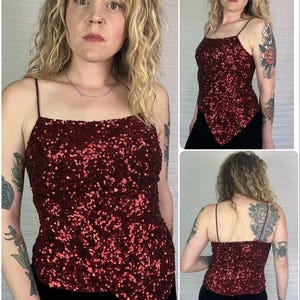 May include: A burgundy sequin camisole top with thin spaghetti straps. The top has a square neckline and a unique asymmetrical hemline. The sequins create a shimmering effect, perfect for a night out. The top is paired with black bottoms.