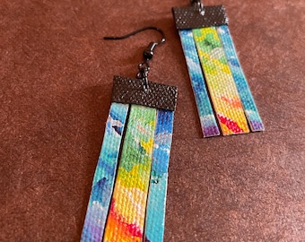 May include: A pair of dangle earrings with a colorful abstract design. The earrings are made of fabric strips with a rainbow color scheme, including blue, green, yellow, orange, and red. The fabric strips are attached to a black metal hook.