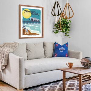 May include: A framed illustration of a pier with the text "Newport Beach" in a retro style. The illustration is in a light blue frame and is hanging on a white wall above a light gray couch with a blue and red patterned pillow.