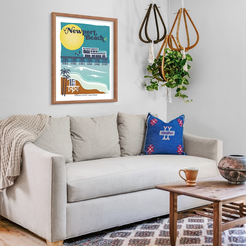 May include: A framed illustration of a pier with the text "Newport Beach" in a retro style. The illustration is in a light blue frame and is hanging on a white wall above a light gray couch with a blue and red patterned pillow.