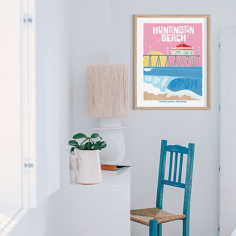 Op de afbeelding: Een ingelijste kunstprint met een kleurrijke illustratie van Huntington Beach, Californi&euml;. Het kunstwerk toont een pier, oceaangolven en de tekst "Huntington Beach" in wit tegen een roze achtergrond. De print zit in een houten lijst.