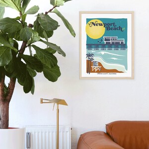 May include: A retro-style illustration of the Newport Beach pier with a lifeguard stand, palm trees, and a yellow sun. The text "Newport Beach" is in a stylized font. The image is framed in a light wood frame.