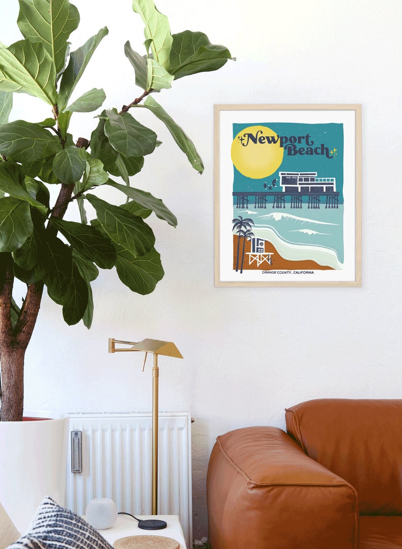 May include: A retro-style illustration of the Newport Beach pier with a lifeguard stand, palm trees, and a yellow sun. The text "Newport Beach" is in a stylized font. The image is framed in a light wood frame.