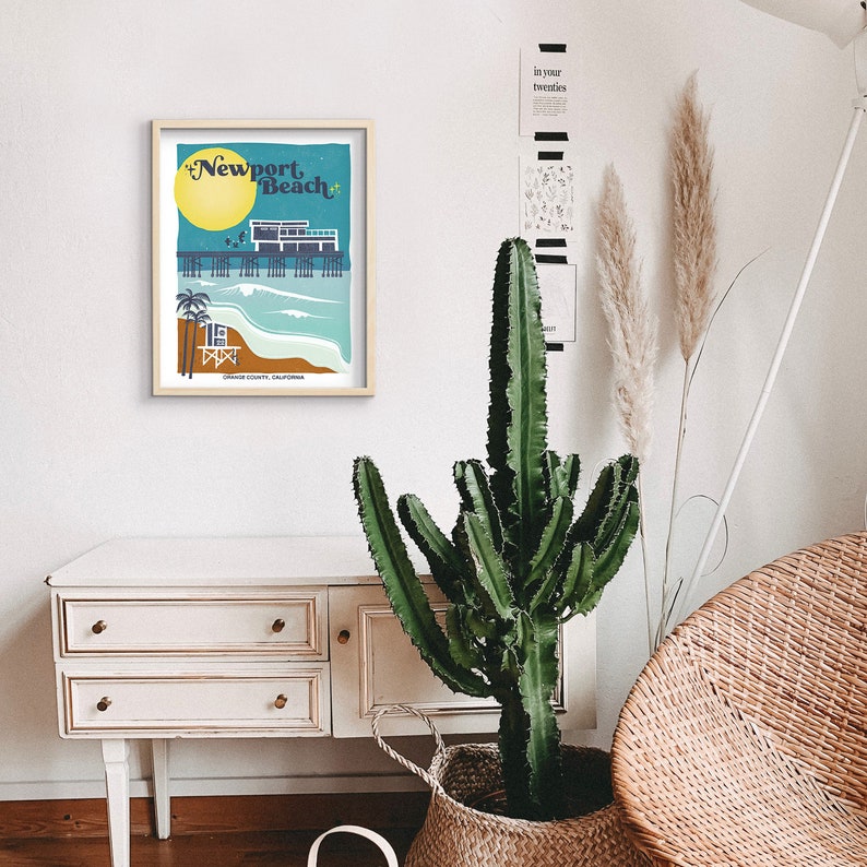 May include: A framed illustration of the Newport Beach pier with a blue and green color scheme. The text "Newport Beach" is in a retro font. The illustration is framed in a light wood frame and is hanging on a white wall.