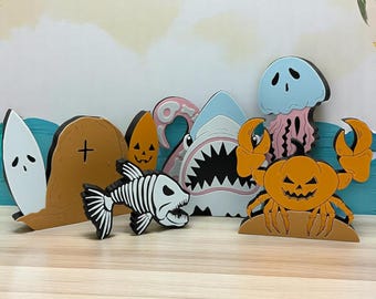 May include: A set of seven colorful wooden Halloween-themed ocean creature figurines. The set includes a shark, a crab, a jellyfish, a ghost, a skeleton fish, a tombstone, and a pumpkin.