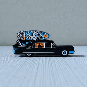 May include: A black hearse-shaped object with a surfboard on top. The surfboard has a blue, orange, and white design with skulls and waves. The hearse has blue and orange accents and white wheels.
