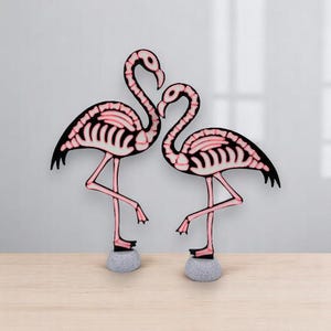 May include: Two decorative flamingo sculptures. The flamingos are black with pink skeleton detailing and pink legs. They stand on gray stone bases. The flamingos are posed facing each other.