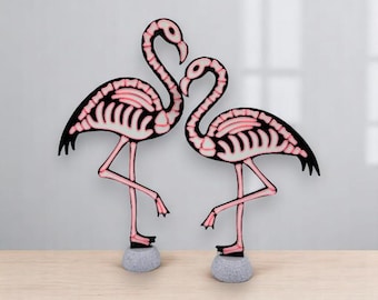 May include: Two decorative flamingo sculptures. The flamingos are black with pink skeleton detailing and pink legs. They stand on gray stone bases. The flamingos are posed facing each other.