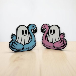 May include: Two enamel pins featuring a ghost riding a flamingo float. One pin is blue and the other is pink.