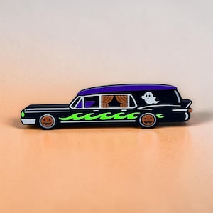 May include: A black enamel pin shaped like a hearse with a purple roof, bright green flames along the side, and a white ghost graphic. The wheels feature orange jack-o'-lantern faces, and the side windows show tan curtains inside the vehicle.