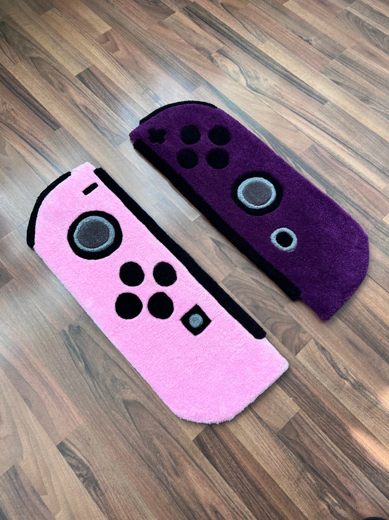 May include: Two plush rugs shaped like Nintendo Switch Joy-Con controllers. One rug is pink and the other is purple.