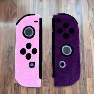 May include: Two plush rugs shaped like Nintendo Switch controllers. The left rug is pink with black buttons and the right rug is purple with black buttons.