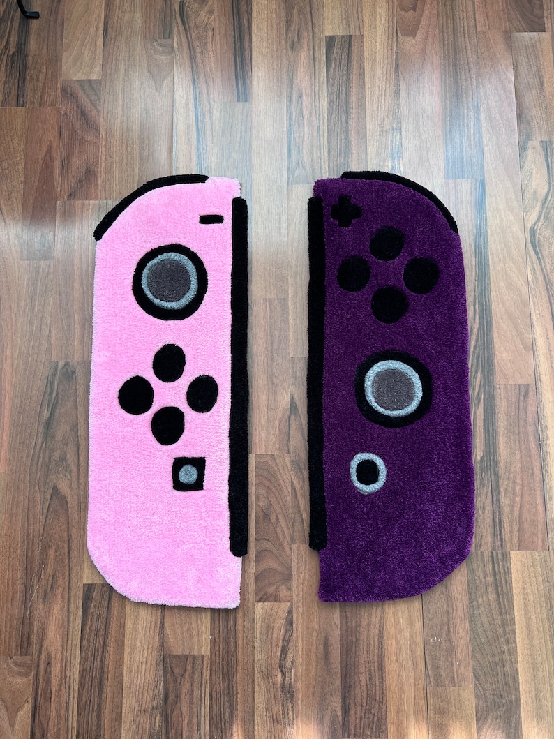 May include: Two plush rugs shaped like Nintendo Switch controllers. The left rug is pink with black buttons and the right rug is purple with black buttons.