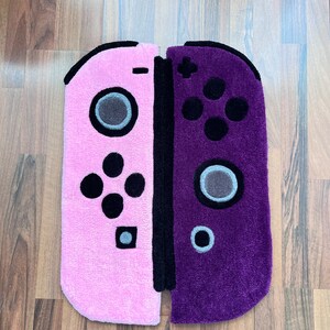 May include: A pink and purple rug shaped like a Nintendo Switch game console. The rug is made of soft, plush material and features a detailed design of the console's buttons and controls.